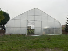 A large arched greenhouse with a transparent plastic cover and metal frame, situated outdoors on a grassy field under an overcast sky.