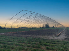 A large metal hoop house frame structure extending over a prepared dirt field in an open rural landscape under a clear blue sky at sunset.