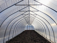 A long greenhouse tunnel with a translucent plastic roof and metal frame, showing dark soil covered beds along the floor.